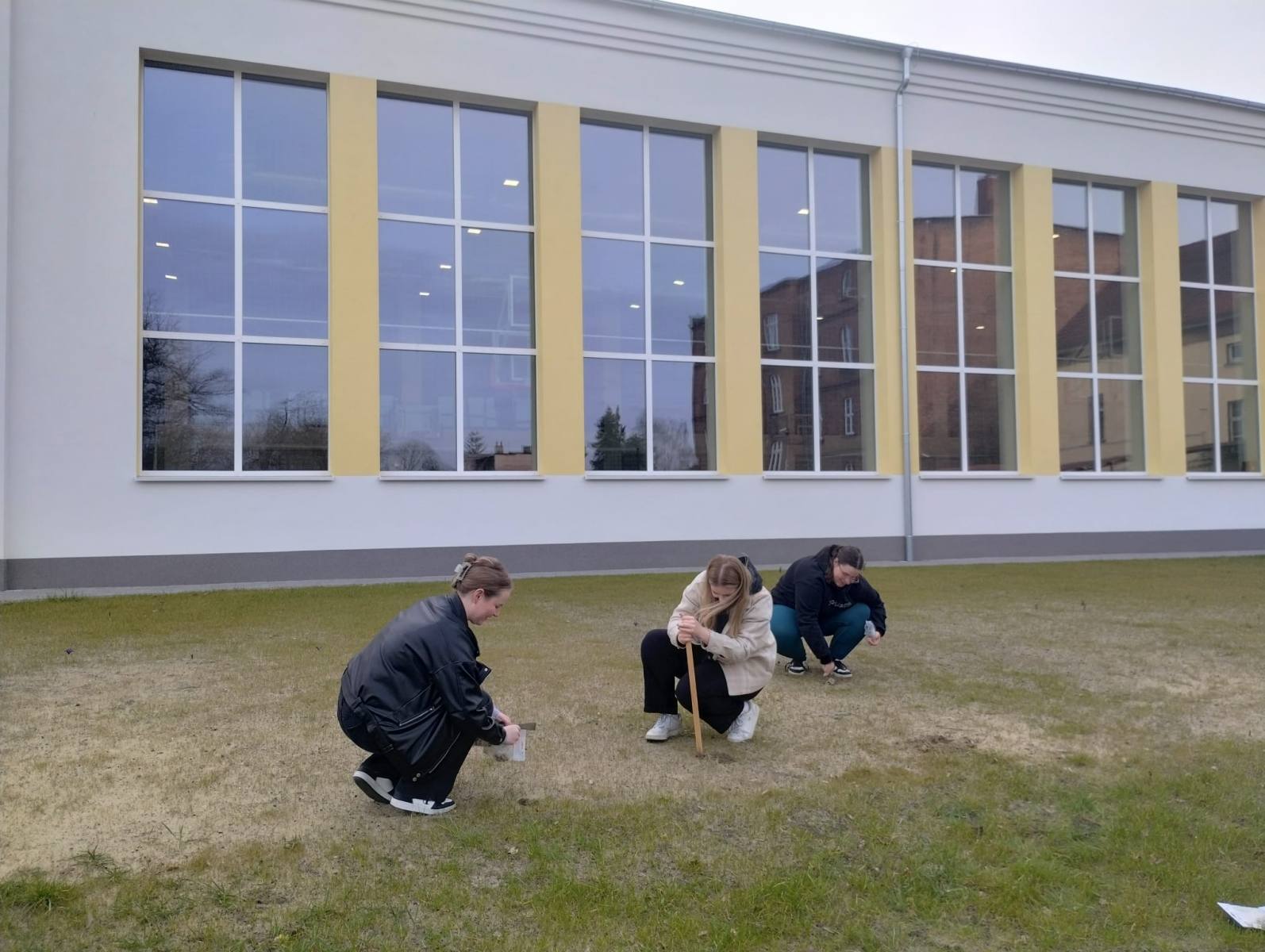 Na zdjęciu dwie uczennice sadzą cebulki kwiatów, jedna uczennica robi dołek w ziemi. W tle sala gimnastyczna.