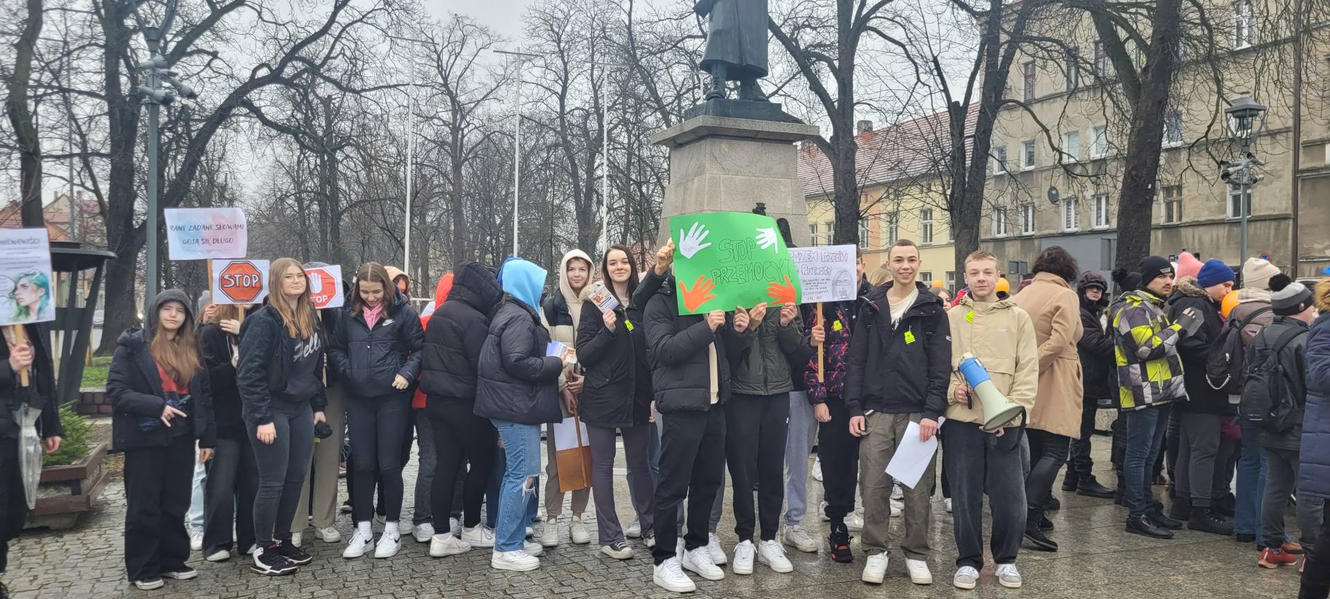 uczniowie stoją w rzędzie trzymając w rękach banery, plakaty oraz megafon. Uśmiechają się. W tle pomnik żołnierza.