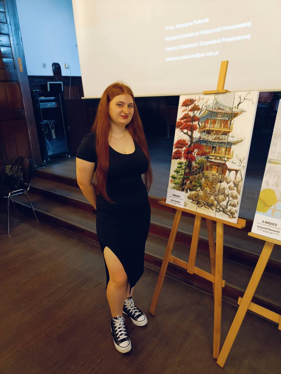Dziewczyna ubrana w czarną sukienkę stoi obok sztalugi z obrazem.