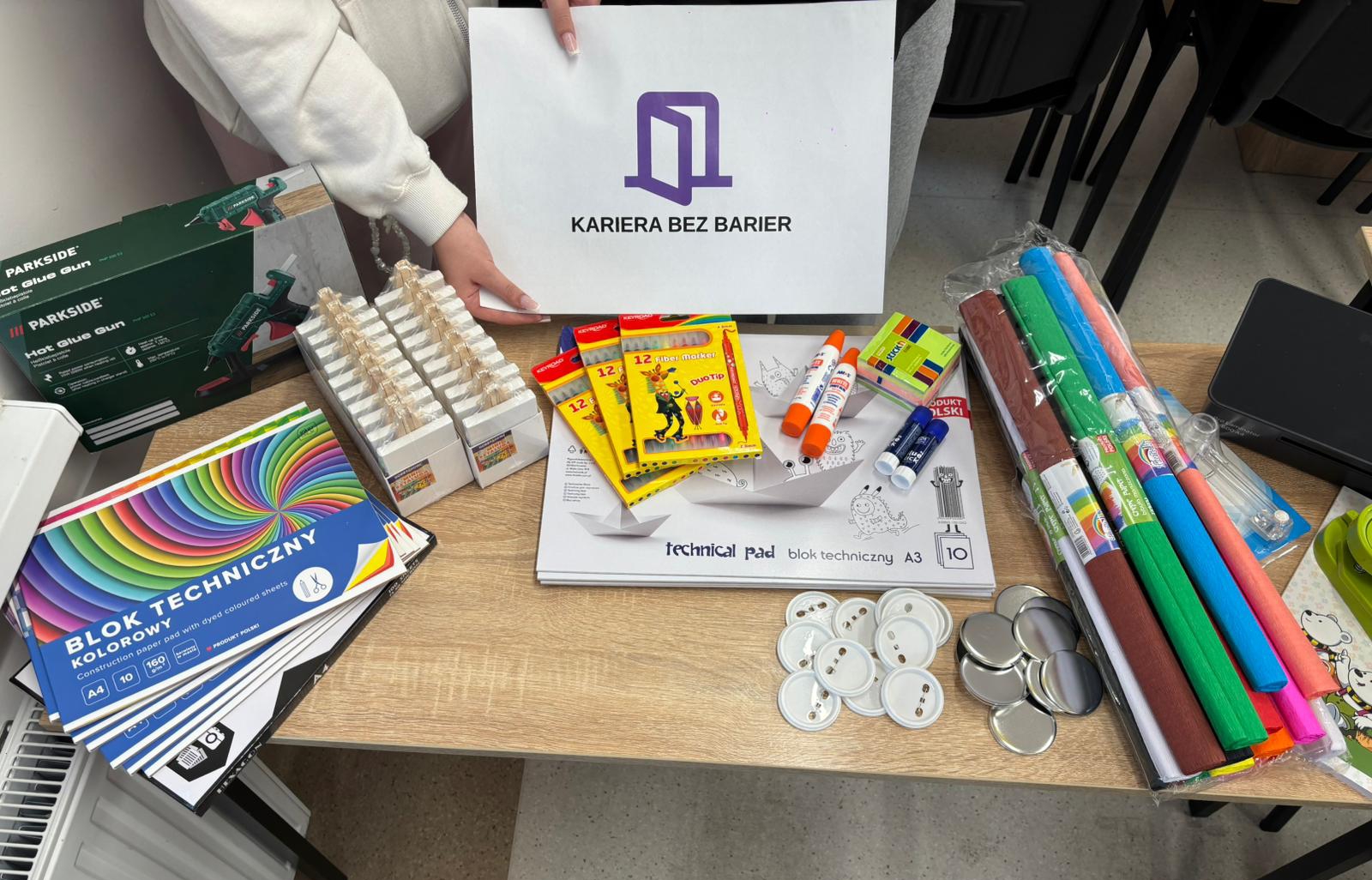 Zdjęcie przedstawia uczniów, którzy prezentują materiały papiernicze, laminator oraz maszynkę do przypinek.